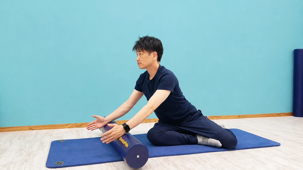 男性がピラティスマットの上でポーズをとっている。フォームローラーを体の前に横に置き、手のひらを向かい合わせにして乗せている。右脚を前、左足を後ろにして膝を曲げて座っている。